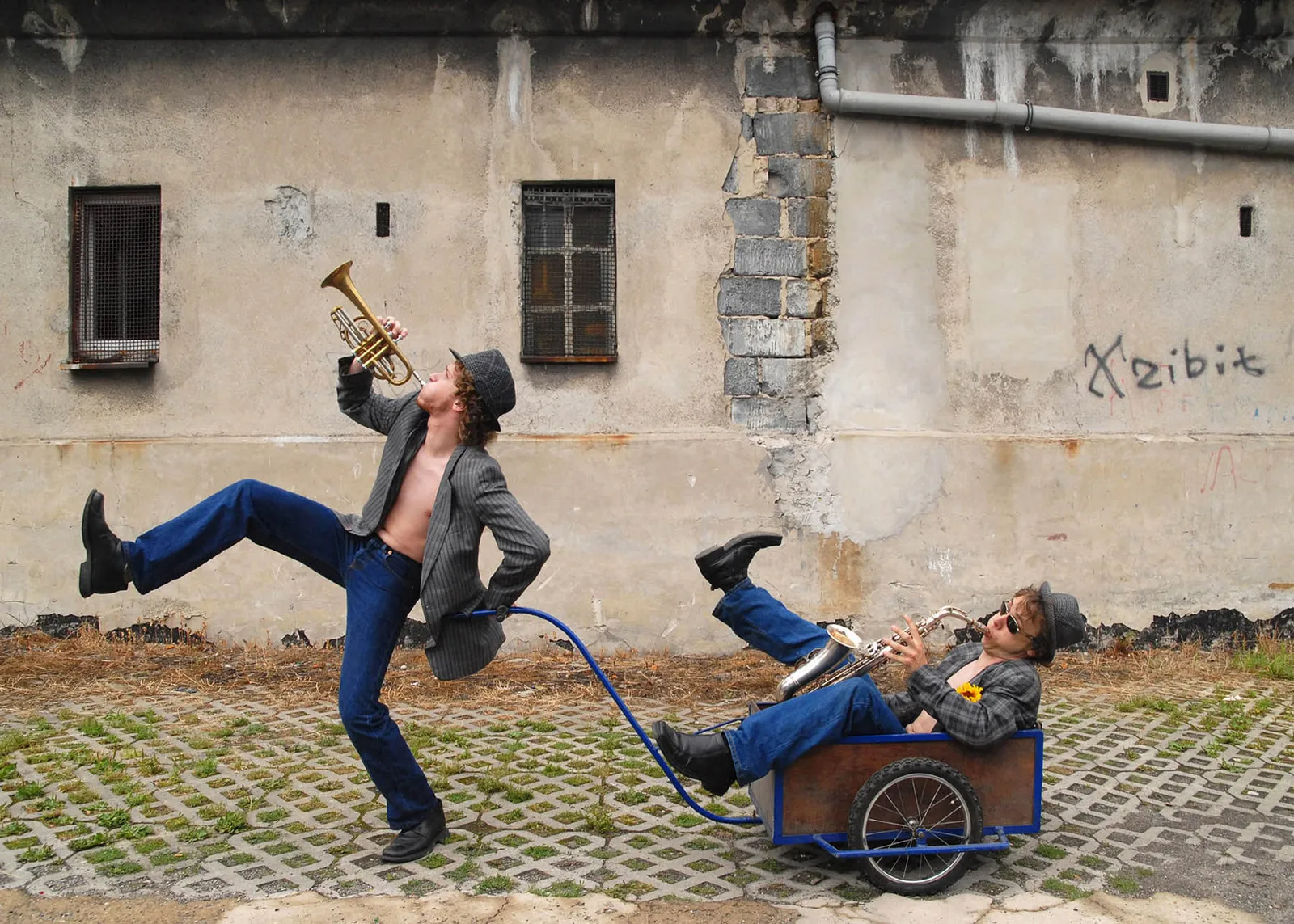 Two musicians in a surreal street scene, connected by a blue hose, playing trumpet and saxophone against a derelict wall