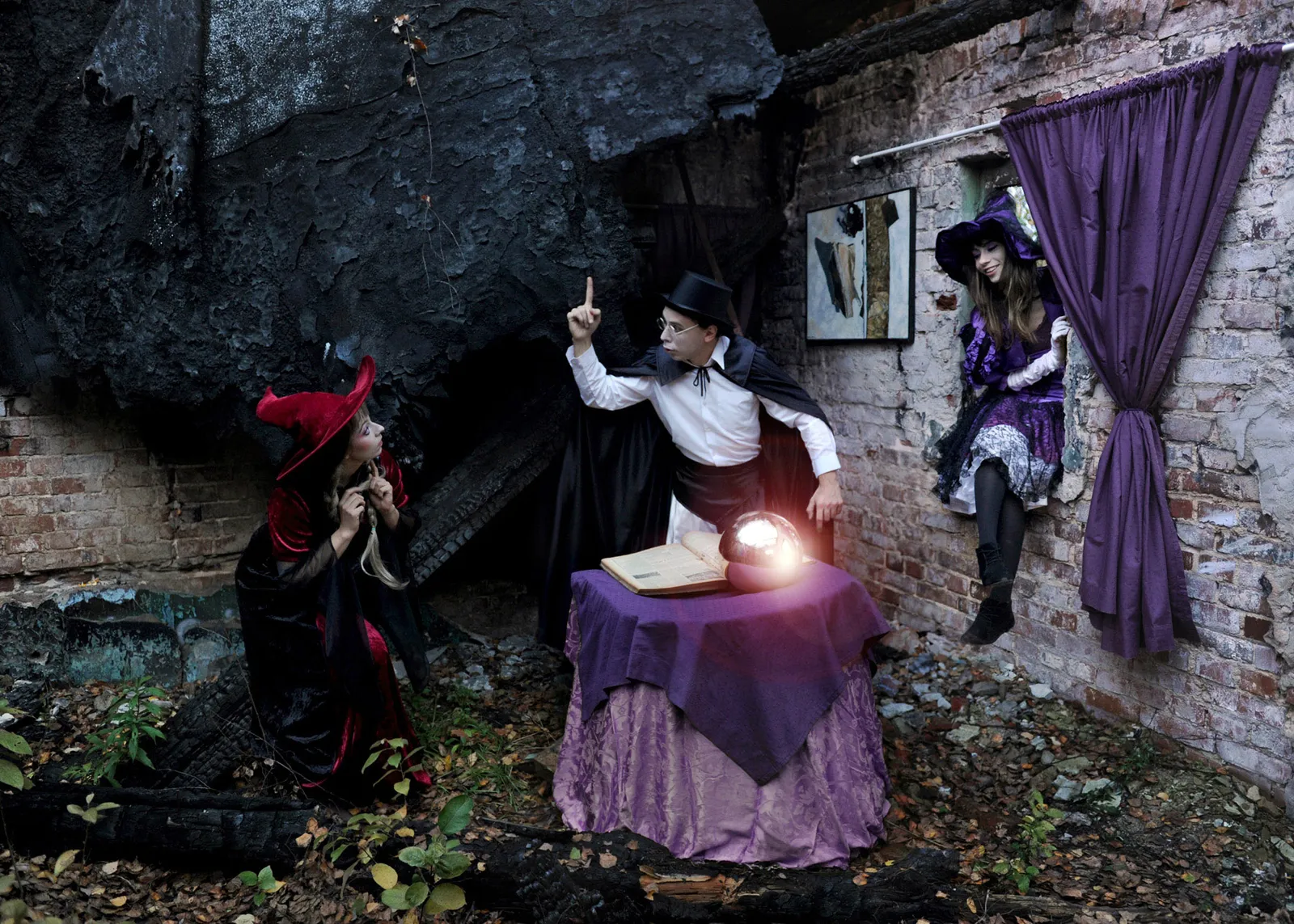 Three figures dressed as witches and a magician gathered around a glowing orb on a table, in a ruined interior with a purple curtain