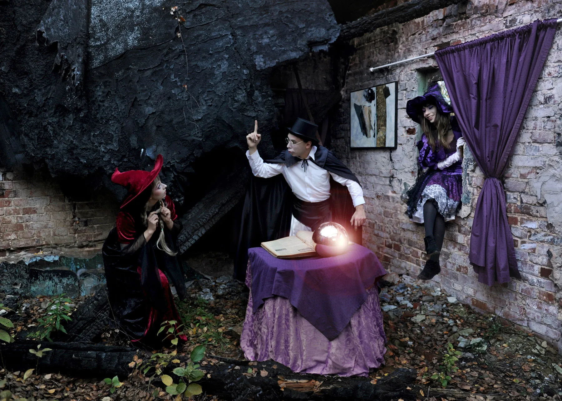 Three figures dressed as witches and a magician gathered around a glowing orb on a table, in a ruined interior with a purple curtain