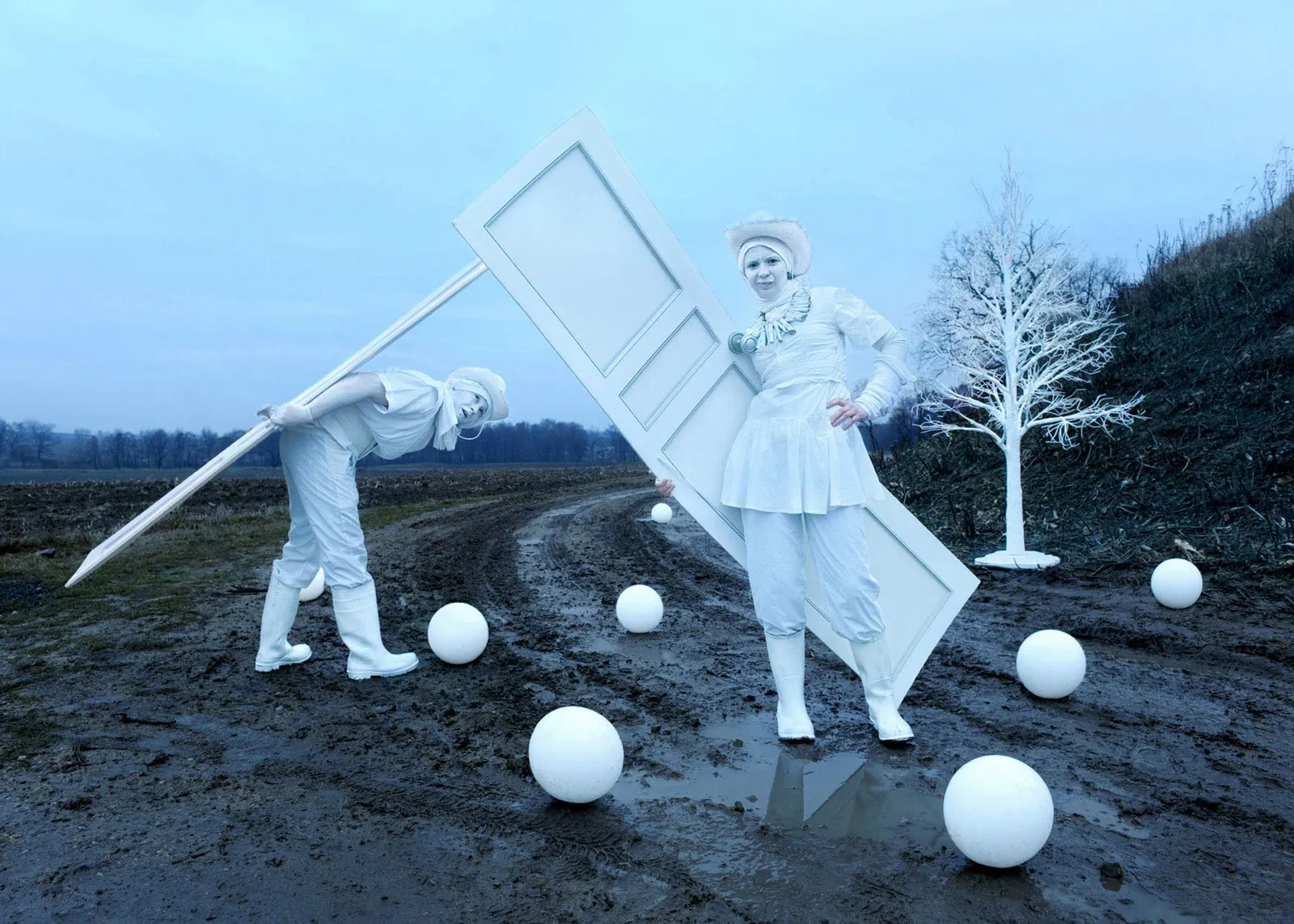 Two figures dressed entirely in white carry an oversized white door across a muddy field, surrounded by white spheres
