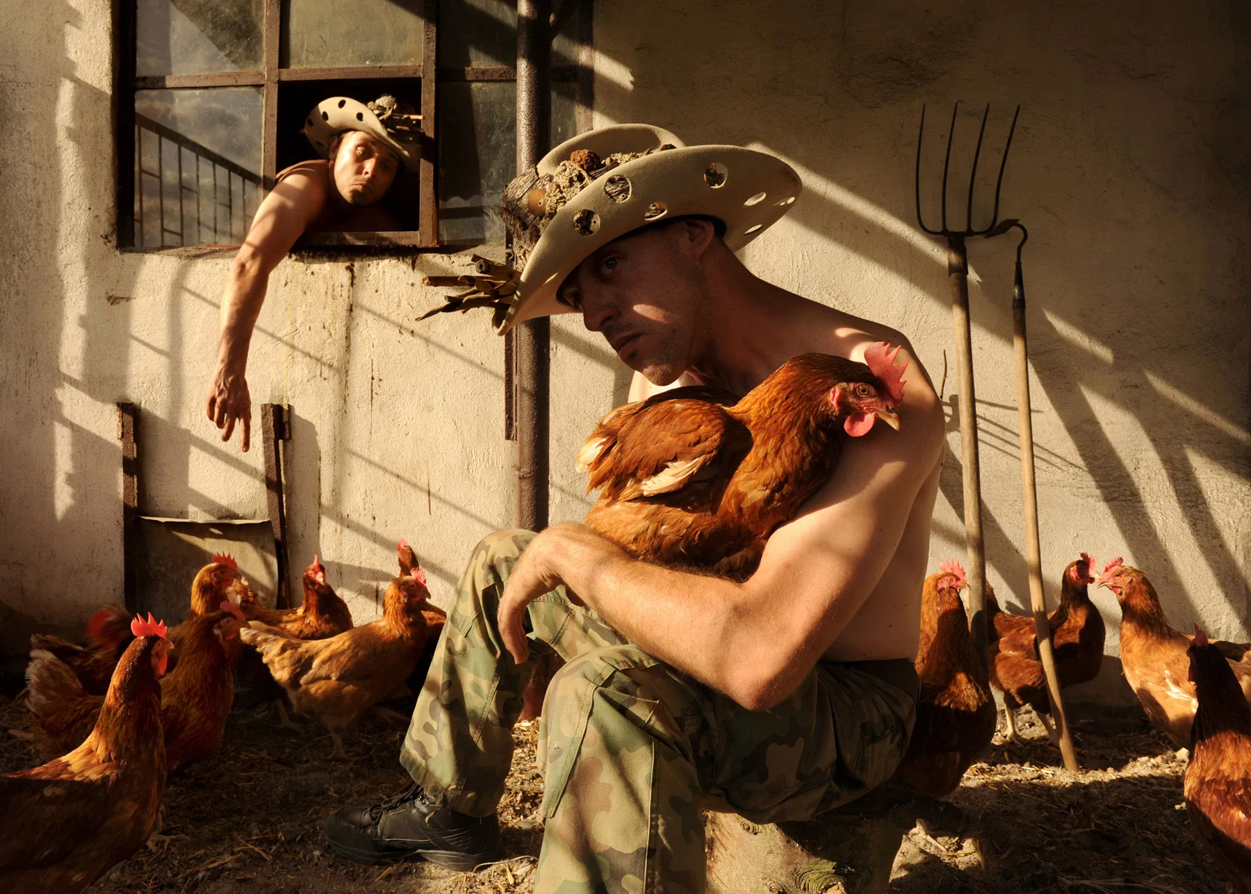 A man in a makeshift hat holds a hen in a farmyard, another figure leans out of a window nearby