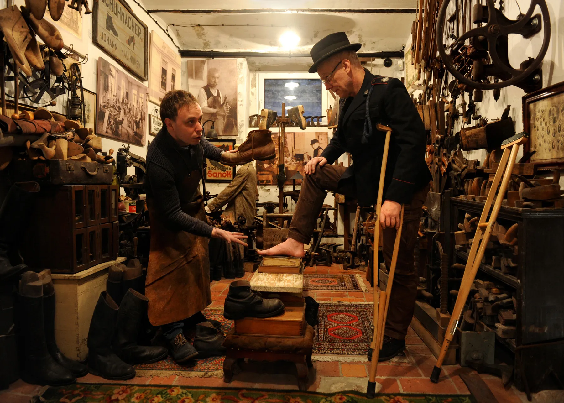 A scene in a shoemaker's workshop — one man fitting a boot on another, surrounded by antique cobbler's tools