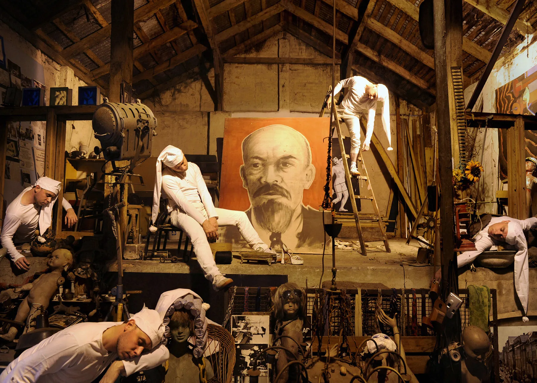 Figures in white inside an old barn beneath a large orange portrait, surrounded by discarded mannequins and dolls