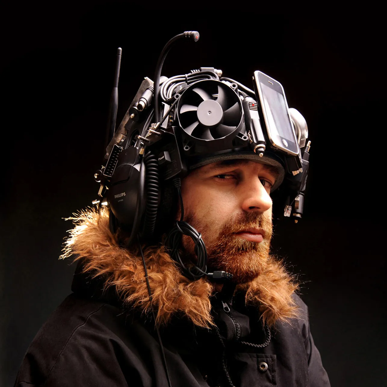 Portrait of a bearded man in a fur-trimmed parka, wearing a helmet built from electronics — a fan, a phone, and headphones